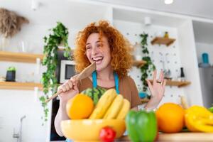 Funny beautiful woman singing into spatula, cooking in modern kitchen, holding spatula as microphone, dancing, listening to music, playful girl having fun with kitchenware, preparing food. photo