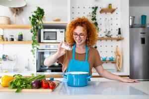 Cute young woman cooking and adding spice to meal, laughing and spending time in the kitchen photo