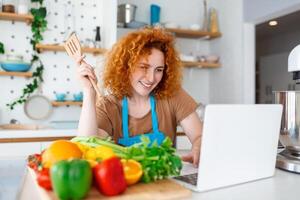 un joven mujer aprende a cocinar, ella relojes vídeo recetas en un ordenador portátil en el cocina y cocinar un plato . Cocinando a hogar concepto foto