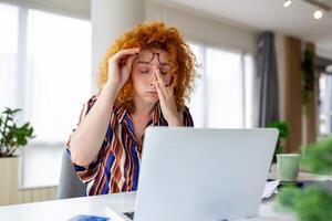 Exhausted businesswoman having a headache in her office. Overworked businesswoman experiencing work fatigue while siting in front of a laptop. photo