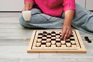 mujer hace un moverse en un juego de juego de damas a hogar mientras sentado en el piso y participación un jarra de té foto