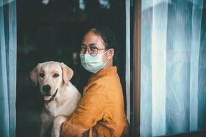 asiático niña vistiendo anteojos, vistiendo un mascarilla, con Labrador perro mirando fuera el ventana, es aburrido de teniendo a detener y tratar el enfermedad a hogar solo. concepto hogar cuarentena, prevención covid-19 foto