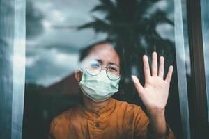 asiático niña vistiendo anteojos, vistiendo un mascarilla, mirando fuera el ventana, es aburrido de teniendo a detener y tratar el enfermedad a hogar solo. concepto hogar cuarentena, prevención covid-19 foto