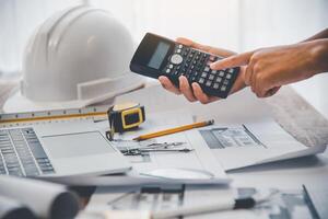 ingeniero utilizar calculadora trabajando en oficina con planos, inspección en lugar de trabajo para arquitectónico plan, construcción proyecto, negocio construcción foto