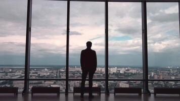 arrière vue de homme dans formel suites qui supporter dans de face de panoramique fenêtre avec ville voir. une homme des stands dans de face de gros panoramique fenêtre vue de dos. video