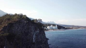 video. aéreo ver de hotel en mar apuntalar y montañas cubierto con verde bosque, arboles con azul cielo a amanecer. video. aéreo ver en costa con lujo hotel con bosque y montañas antecedentes video