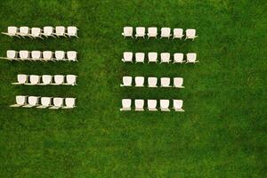 Top view of White wooden chairs standing in rows on a green field waiting for guests.View from a height of many white chairs standing on a green lawn photo