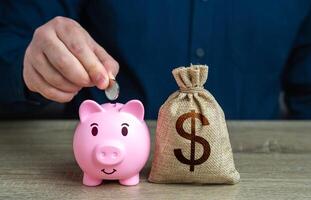 A man puts coins in a piggy bank near dollar money bag. Accumulating money. Savings and investments. Collection of donations. Bank deposit. Raise funds for your dream. Reduce costs. photo