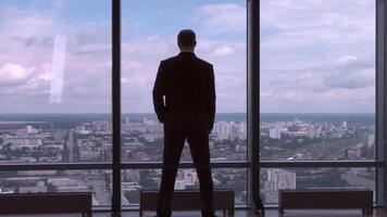 Full length back view of successful businessman in suit standing in office with hands on waist, CEO. Businessman from the back in front of a city view on the window video