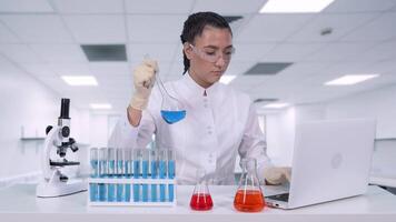 un joven hembra investigador análisis un azul líquido en un matraz y lo hace clínico ensayos. un hembra científico conduce genético investigación y escribe el resultados a un ordenador portátil en un innovación laboratorio video