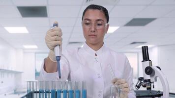 uma jovem fêmea cientista dentro branco laboratório casaco protetora óculos e luvas examina uma azul líquido usando uma micropipeta e teste tubo. uma fêmea investigador é conduzindo clínico ensaios. Ciência laboratório video