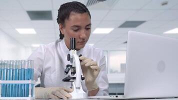 A young female researcher in a white coat looks at biological samples under a microscope, conducts clinical trials and writes data to a laptop while sitting at a white table. Scientific laboratory video