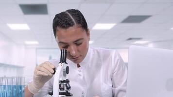 A young female medical professional in a white coat looks at biological samples under a microscope and writes data to a laptop while sitting at a white table. Scientific laboratory video