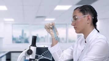 joven hembra laboratorio técnico en un blanco Saco realiza laboratorio pruebas de azul líquido en un prueba tubo mientras sentado a un mesa en un Ciencias laboratorio.lento movimiento video