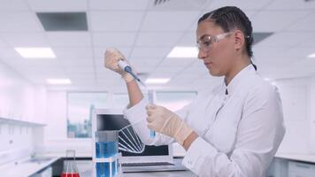 A young female laboratory technician in a white lab coat transfers a sample of a blue liquid into a test tube using a micropipette sitting at a table in a science lab. video