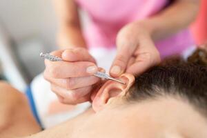 Crop beautician applying flexible massage brass ear pen on female client in salon photo