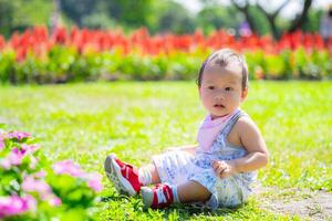 Asian Toddler 1 year old Sitting Amongst Colorful Garden Flowers. Young child sits thoughtfully on grass, surrounded by burst of garden colors, showcasing the innocence and beauty of early childhood. photo