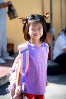 contento niño niña vistiendo reno horquilla y que lleva mochila, Listo para festivo colegio día, sonriente asiático niño con marrón reno banda para el cabello a obtener dentro Navidad festival. foto