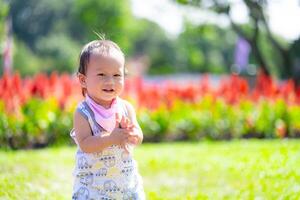 Joyful Toddler Clapping Hands in Flower Garden. Delighted baby boy claps his hands while standing in lush garden, with a backdrop of vivid red flowers and bright greenery. Asian child aged 1 year old. photo