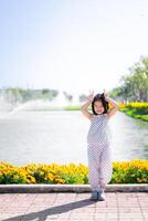 niño juguetón gesto por el orilla del lago jardín, joven niña en lunares atuendo soportes por orilla del lago jardín, haciendo juguetón paz firmar con su dedos, radiante felicidad en brillante al aire libre ajuste. foto