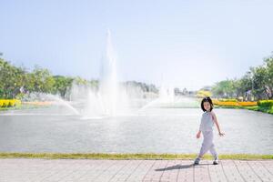 Asian Girl Strolling by Park Fountain on Sunny Day, Kid in dotted outfit takes leisurely stroll by majestic fountain in the park, with sun casting a soft glow on serene landscape. Child aged 6 years. photo