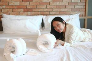 A woman in a white dress is lounging in the bedroom on a white bed, looking at the camera. photo