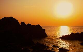 The sun is setting on the sea with rocks in the foreground, black silhouettes on the rocks, light reflecting off the sea. photo