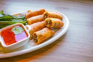 Freshly fried golden-yellow spring rolls are placed on a white plate with various vegetables and a delicious dipping sauce. photo