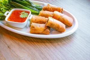 Freshly fried golden-yellow spring rolls are placed on a white plate with various vegetables and a delicious dipping sauce. photo