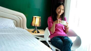 retrato de hermosa joven asiático mujer participación taza de Mañana café en blanco dormitorio, feliz, alegre, relajante en verano. tailandés asiático maquillaje ideas, blanco dormitorio foto