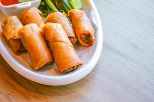 recién frito amarillo dorado primavera rollos son metido en un blanco plato con varios vegetales y un delicioso inmersión salsa. foto