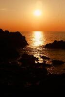 The sun is setting on the sea with rocks in the foreground, black silhouettes on the rocks, light reflecting off the sea. photo