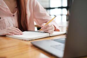 Close-up of a student's hand writing notes in a notebook while engaged in online learning, with a laptop visible. photo