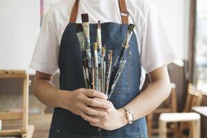 A close-up of the artist hands wearing an apron smeared with paint. Clutching many brushes and paintbrushes. photo