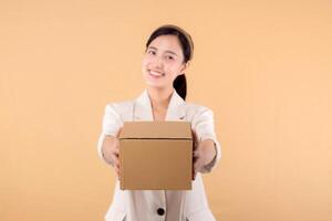 portrait of happy successful confident young asian business woman wearing white jacket holding parcel box isolated on beige studio background, Delivery courier and shipping service concept. photo