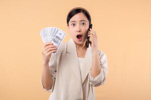 portrait of happy successful confident young asian business woman wearing white jacket holding smartphone and cash money dollar standing over beige background. millionaire business, shopping concept. photo