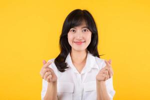 Young female healthcare. Asian woman wearing white shirt feels happy and romantic shapes heart gesture expresses tender feeling poses isolated on yellow background. People affection and care concept. photo