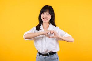 joven hembra cuidado de la salud. asiático mujer vistiendo blanco camisa siente contento y romántico formas corazón gesto expresa oferta sensación poses aislado en amarillo antecedentes. personas afecto y cuidado concepto. foto