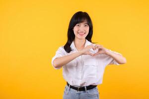 Young female healthcare. Asian woman wearing white shirt feels happy and romantic shapes heart gesture expresses tender feeling poses isolated on yellow background. People affection and care concept. photo