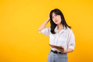 Doubtful Asian young adult business woman working on digital tablet and looking away thinking of solution isolated on yellow background, considering offer, making decision or feeling doubt concept. photo