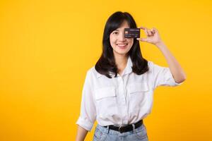 asiático joven mujer participación crédito tarjeta en frente de uno ojo con contento sonrisa aislado en amarillo antecedentes. pago compras en línea concepto. foto
