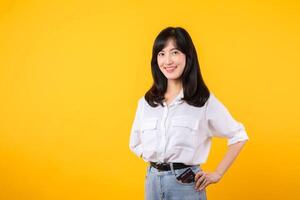 young fun woman of Asian ethnicity wear white shirt and denim jean hold put or pull credit bank card from pocket isolated on yellow background. Lifestyle online payment shopping concept. photo