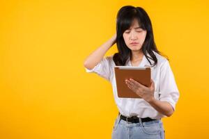Doubtful Asian young adult business woman working on digital tablet and looking away thinking of solution isolated on yellow background, considering offer, making decision or feeling doubt concept. photo