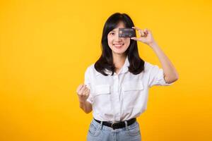 Asian young woman holding credit card in front of one eye and showing fist hand gesture with happy smile isolated on yellow background. Payment shopping online concept. photo