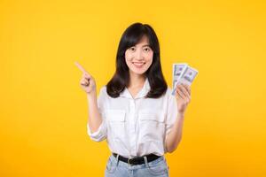 Young happy woman of Asian ethnicity wear white shirt and denim jean holding cash money in dollar and pointing finger to free copy space against yellow background. Investment and financial concept. photo