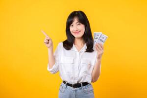 Young happy woman of Asian ethnicity wear white shirt and denim jean holding cash money in dollar and pointing finger to free copy space against yellow background. Investment and financial concept. photo