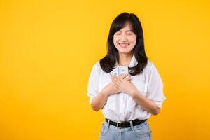 Happy young asian businesswoman wearing white shirt and denim jean holding cash, hugging dollars money and smiling, standing over yellow background. Love to be rich concept. photo