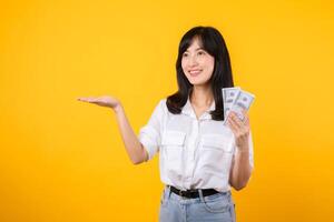 Young happy woman of Asian ethnicity wear white shirt and denim jean holding cash money in dollar and pointing hand to free copy space against yellow background. Investment and financial concept. photo