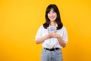 Happy young asian businesswoman wearing white shirt and denim jean holding cash, hugging dollars money and smiling, standing over yellow background. Love to be rich concept. photo