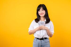 Happy young asian businesswoman wearing white shirt and denim jean holding cash, hugging dollars money and smiling, standing over yellow background. Love to be rich concept. photo